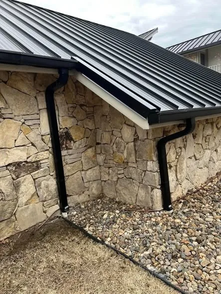 Black gutters and downspouts at the corner of a stone building with a metal roof